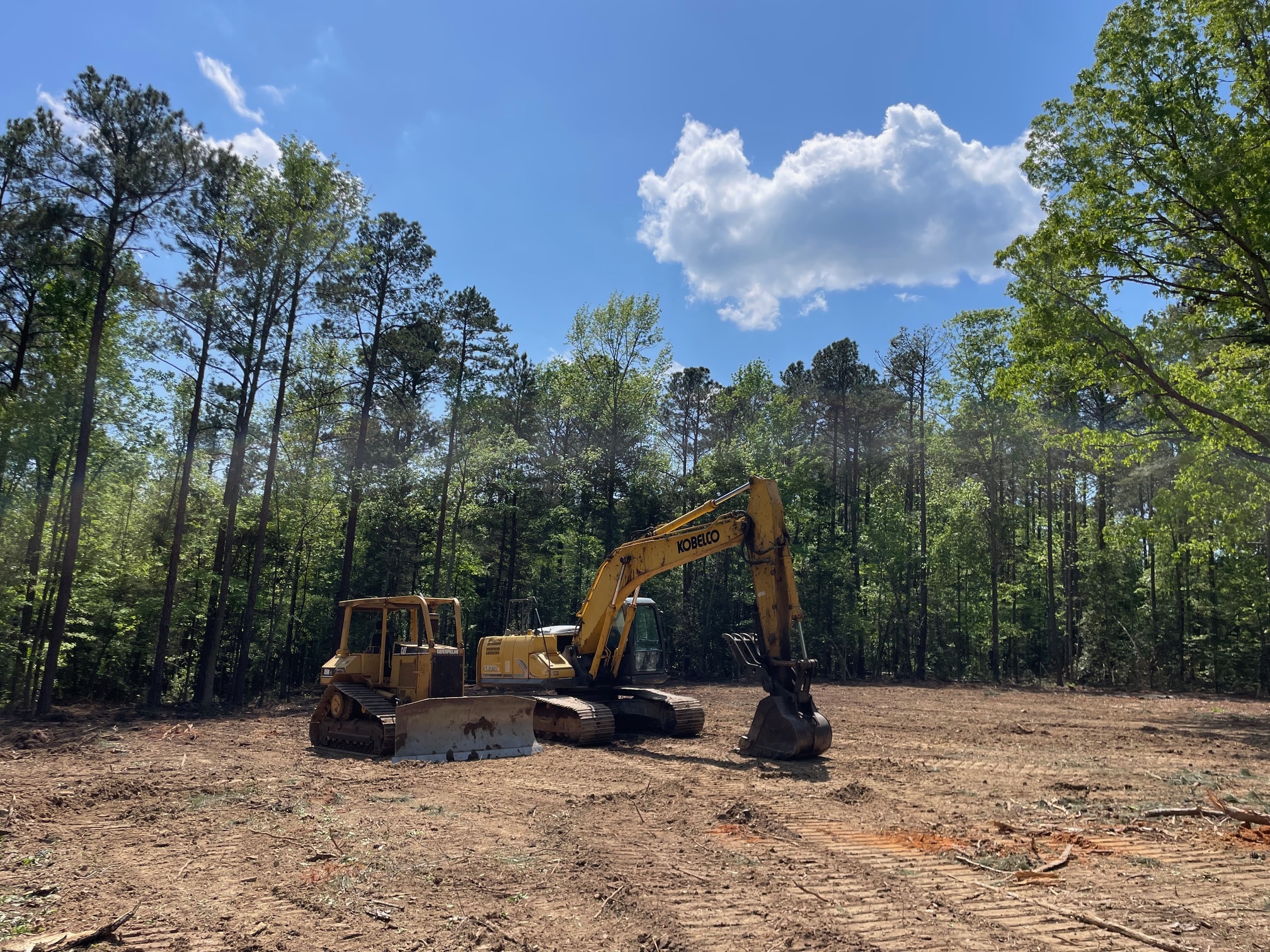 Bulldozer and Kobelco excavator parked on a cleared plot in a wooded area under blue sky