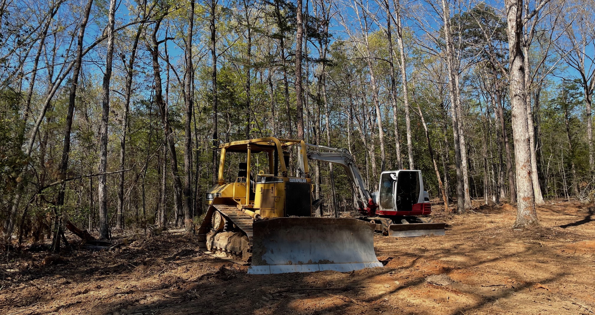 Bulldozer and compact excavator parked on a cleared forest lot under blue sky